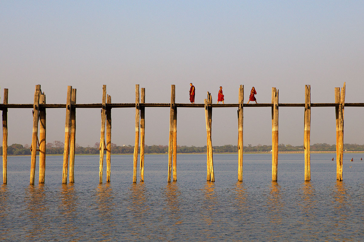 Red robed Buddhist monks walk across teak bridge